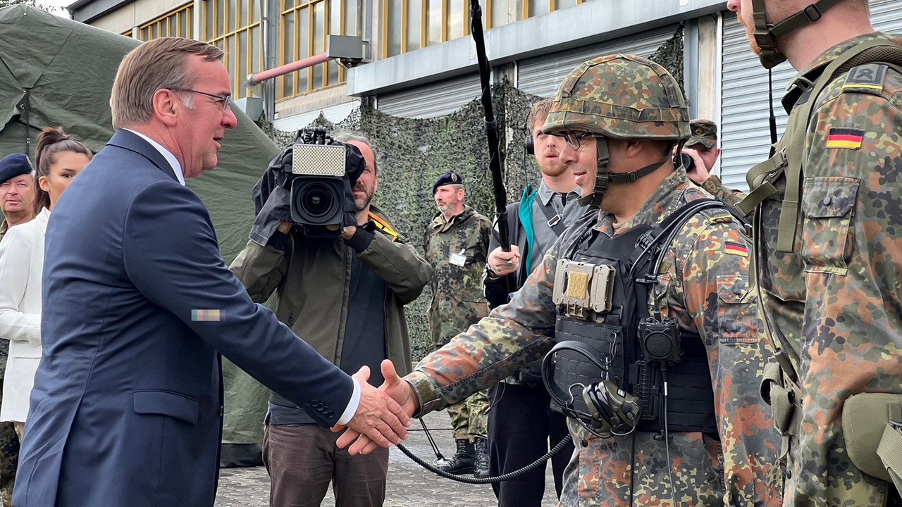 Zu sehen ist der Bundesverteidigungsminister Boris Pistorius, wie er einem Soldaten der "Cyber-Truppe", der eine Camouflage-Uniform trägt, die Hand schüttelt.