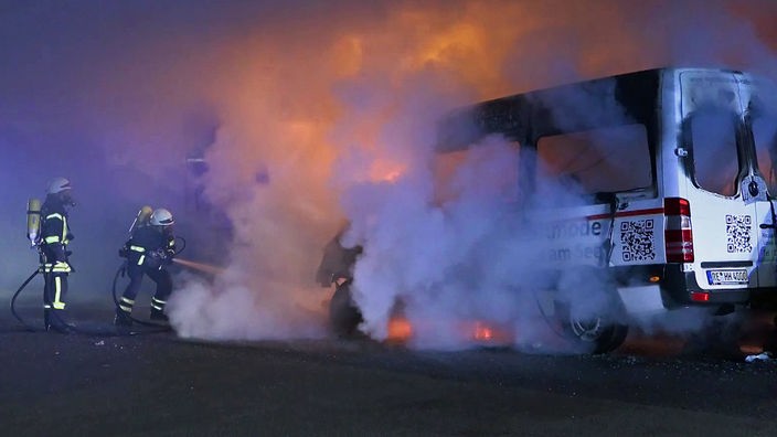 Ein Kleinbus brennt auf einer Straße in Köln-Porz und zwei Feuerwehrleute versuchen, ihn zu löschen