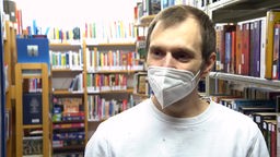 Man sieht einen mann, der eine FFP2 Maske trägt und vor Bücherregalen steht.