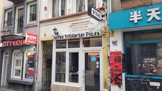 Ein kleiner, verlassener Pizzaladen von außen, nebendran jeweils andere Geschäfte