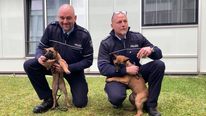 Hundeführer Marc Pfeiffer sitzt mit Welpe Jax auf einer Wiese, daneben Hundeführer Mark Renner mit Thora (v.l.)