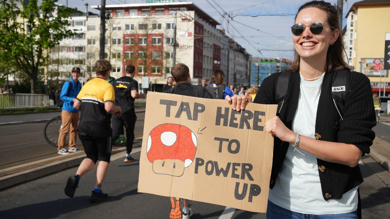 Eine junge Frau steht am Straßenrand und hält ein Pappschild in der Hand. Darauf ist ein Comic-Pilz aus dem Spiel "Mario Kart" zu sehen. Daneben steht: Tap here to power up.