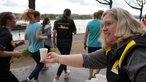 Eine Frau steht an einem Versorgungsstand. Sie reicht in ihrer rechten Hand einen Pappbecher mit Wasser an vorbeilaufende Läufer.