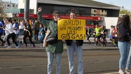 Zwei Frauen stehen am Straßenrand und halten ein gelbes Schild hoch. Darauf steht: Brudi wir müssen los! Gib Gas!