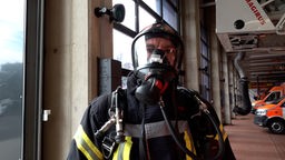 Mann trägt Feuerwehrkleidung und einen Atemschutz, steht in Gerätehalle der Feuerwehr mit Einsatzfahrzeugen im Hintergrund