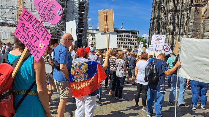 Vor dem Kölner Dom stehen Demonstranten mit Schildern gegen Waffenlieferungen an die Ukraine 