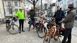 Auf dem Bild sind fünf Menschen zu sehen, welche alle auf oder neben ihrem Fahrrad stehen, während sie sich unterhalten.
