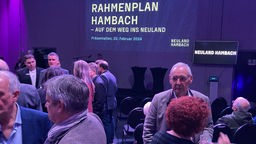 Menschen stehen auf einer Veranstaltung zusammen und networken, im Hintergrund steht "Rahmenplan Hambach - auf dem Weg ins Neuland" an eine Wand projiziert