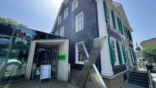 Grau-grünes Schiefer-Fachwerkhaus, im Vordergrund ein großes X aus Metall
