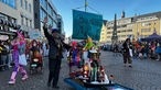 Ein Mann steht neben einem kleinen Wagen, auf dem verschiedene Puppen stehen. Auf einem Schild am Wagen steht "Bonn Alaaf".