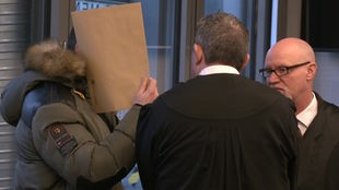 Der Angeklagte steht vor Gericht. Er verdeckt sein Gesicht mit einem Papier.