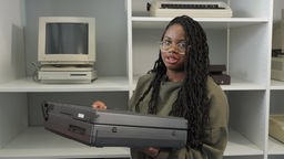 Precious Mutombo hält einen Rechner, hinter ihr ein Regal mit alten Computern