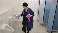 Eine Frau in Putzkleidung mit Lappen und Reinigungsmittel in der Hand auf einer Toilette