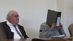 Täter mit Ordner vor dem Gesicht im Gerichtssaal