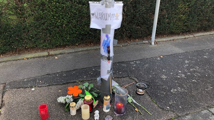 An einem Pfahl an der Straße ist ein Schild mit der Aufschrift "Warum" befestigt, darunter stehen Kerzen und Blumen