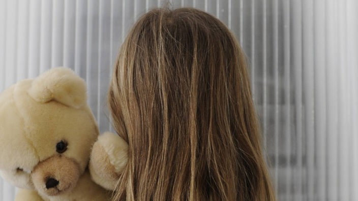 Ein Mädchen mit Teddy auf dem Arm steht vor einer verschlossenen Tür.