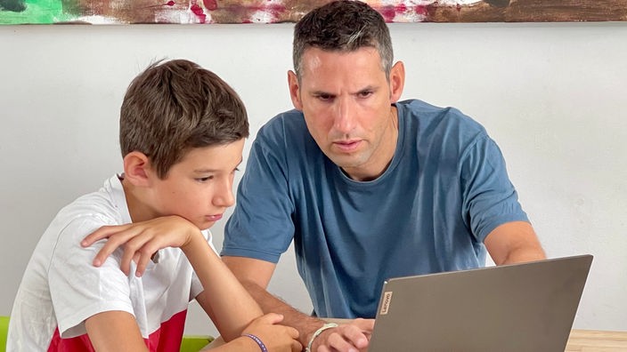 Zu sehen sind ein Vater mit seinem Sohn. Sie sitzen an einem leeren Holztisch und schauen gemeinsam in einen Laptop. 