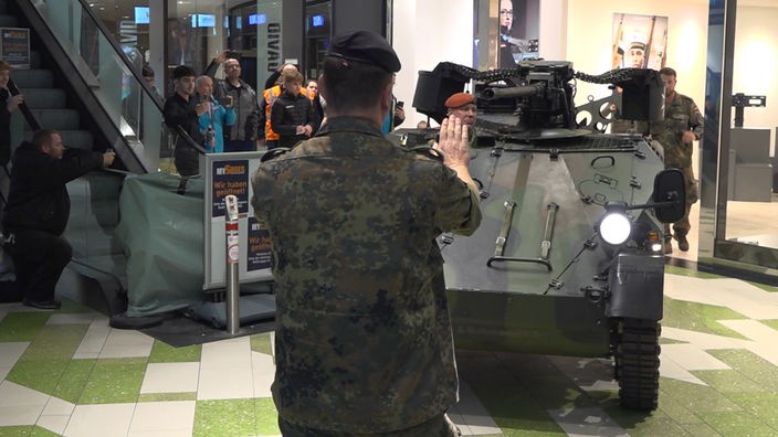 Auf dem Foto ist ein Panzer, der in einem Einkaufszentrum von Soldaten eingewunken wird. An den Seiten stehen Menschen, die sich das anschauen.