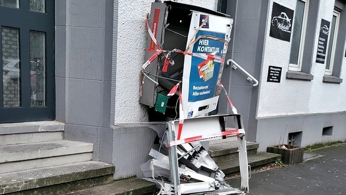 Auf dem Foto ist ein zerbeulter, aufgerissener und mit Absperrband umbundener Zigarettenautomat an einer Hauswand.