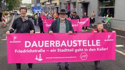 3 Demonstrierende in der Friedrichstraße mit einem Banner im Vordergrund. Auf dem Banner steht: Dauerbaustelle. Friedrichstraße - ein Stadtteil geht in den Keller. Im Hintergrund stehen weitere Demonstrierende mit Bannern.