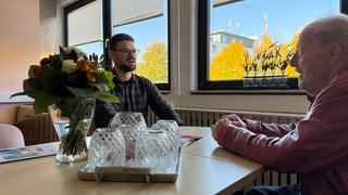 Hans-Walter Ahr sitzt in einer roten Lederjacke gekleidet bei seinem Sozialarbeiter Nathanael Wedler am Tisch, auf dem Tisch stehen Wassergläser und Blumen