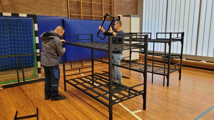 Zwei Männer bauen in einer Sporthalle Stahlgerüste für Etagenbetten auf 