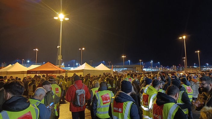 hunderte von Menschen stehen nachts auf dem Flughafengelände. Sie tragen Warnwesten mit Verdi Schriftzug auf rotem Grund.