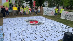 Man sieht Menschen, die mit Transparenten und Bannern in einem Kreis um viele Solidaritätsbekundungen auf Papier, die auf dem Boden liegen, stehen. Es sind streikende Pflegekräfte in Bonn.