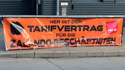 Ein großes orangenes Banner mit der Forderung nach einem Tarifvertrag ist zwischen zwei Pollern befestigt