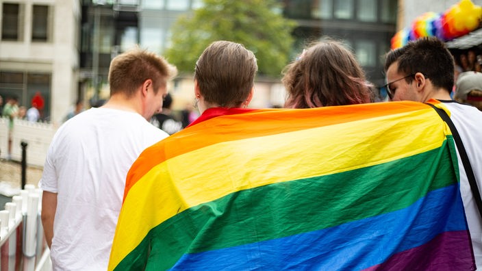 Drei Personen gehen nebeneinander her und tragen auf ihrem Rücken zusammen eine Regenbogenfahne.