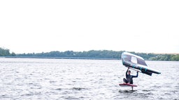 Auf dem Foto ist ein Mann, der mit einem Tragflügel unter dem seinem Surfboard und einem Segel in der Hand surft.