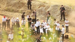 Aktivisten und Musiker, die mehrheitlich weiße Oberteile und schwarze Hosen tragen, stehen auf sandigem Untergrund in der Sonne und werden von Polizisten umstellt.