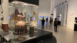 Blick in die Ausstellungshalle: Im Hintergrund laufen Menschen umher, im Vordergrund ist eine Vitrine mit einem ausgestellten Kleid