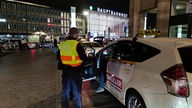 Ein Beamter des Zolls steht vor einem Taxi am Kölner Hauptbahnhof und führt eine Kontrolle durch