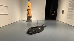 Künstlerische Installation, welche einen "Toten Mann" zeigt, der auf dem Boden liegt und eine Tüte über den Kopf gestülpt hat