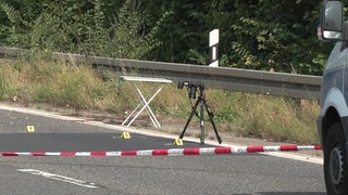 Ein weißer Klapptisch und ein Kamerastativ von der Polizei stehen auf einer Bundesstraße hinte einer Absperrung.