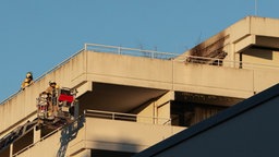 Balkon mit schwarzem Ruß an Fenster. Darunter Feuerwehrleute auf Drehleiter