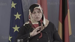 Eine ältere Frau mit Kopftuch steht an einem Pult mit Mikrofonen. Im Hintergrund hängen die europäische, türkische und deutsche Flagge.