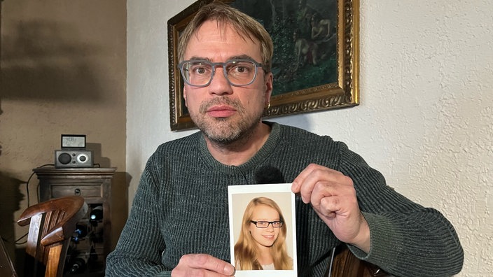 Ein Mann hält ein Foto in der Hand und hält es in die Kamera, darauf zu sehen ist eine junge blonde Frau 