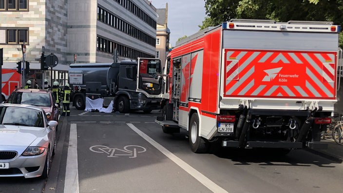 Ein Feuerwehrauto steht auf einer Straße in Köln vor dem Tanklaster, der mit einer Radfahrerin zusammengestoßen ist