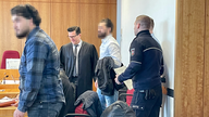 Ein Gerichtssaal. Ein Mann mit einem blau-schwarz karrierten Hemd steht an einem Tisch. Im hinteren Bereich des Bildes kommt ein Mann mit weißem Hemd in den Gerichtssaal. Er wird begleitet von einem Anwalt der eine Robe anhat und einen Polizeibeamten.