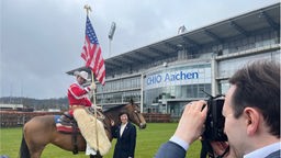 Ein reiter mit Cowboyhut hält eine US Flagge , neben dem Pferd steht eine Frau, im Hintergrund ein Gebäude auf dem "CHIO Aachen" steht