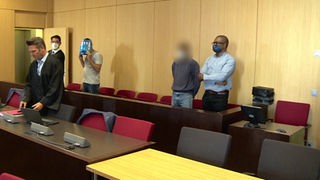 Die zwei mutmaßlichen Täter stehen im Gerichtssaal. Ihr Gesicht ist unerkenntlich. Der Mann links hält sich eine Zeitschrift vor das Gesicht.