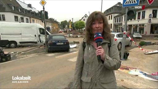 Eine Frau mit gewelltem langen brauen Haar und einem blauen WDR-Mikrofon steht an einer Kreuzung mit demolierten Autos
