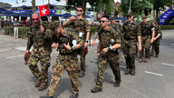 Mehrere Soldaten marschieren, einer von ihnen zeigt die Flagge der Schweiz