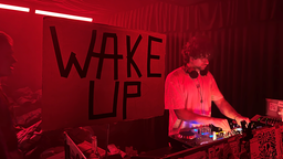 Ein DJ steht vor seinem DJ-Pult, hinter ihm ein großes Schild mit der Aufschritt "Wake Up", das Ganze ist in rotes Licht getaucht