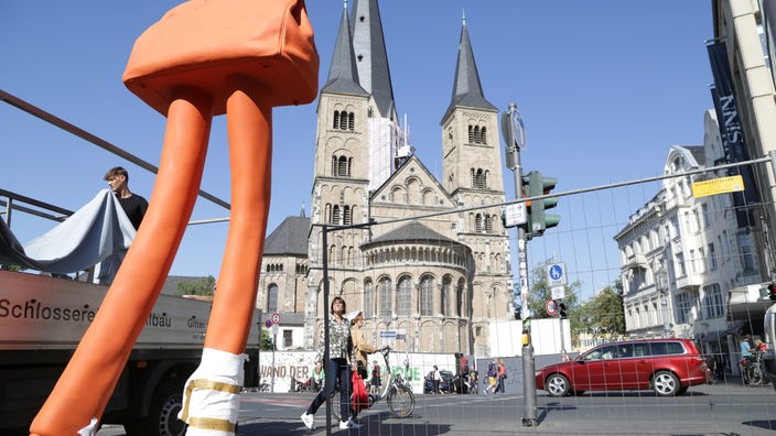 Zu sehen ist die Skulptur einer orangenen Tasche auf sehr langen Beinen - sie steht vor dem Bonner Münster.