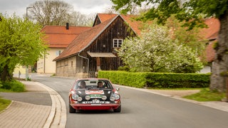 Zwei ältere Männer machen in einem rot-schwarzen alten Porsche-Sportwagen eine Rundfahrt durch ein kleines Dorf 