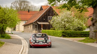 Zwei ältere Männer machen in einem rot-schwarzen alten Porsche-Sportwagen eine Rundfahrt durch ein kleines Dorf 