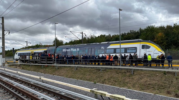 Circa 50 Leute stehen Schlange auf einem Bahnsteig unter freiem Himmel, um in einen weiß-blau-gelben Zug mit der großen Aufschrift "Mireo Plus H" zu gelangen